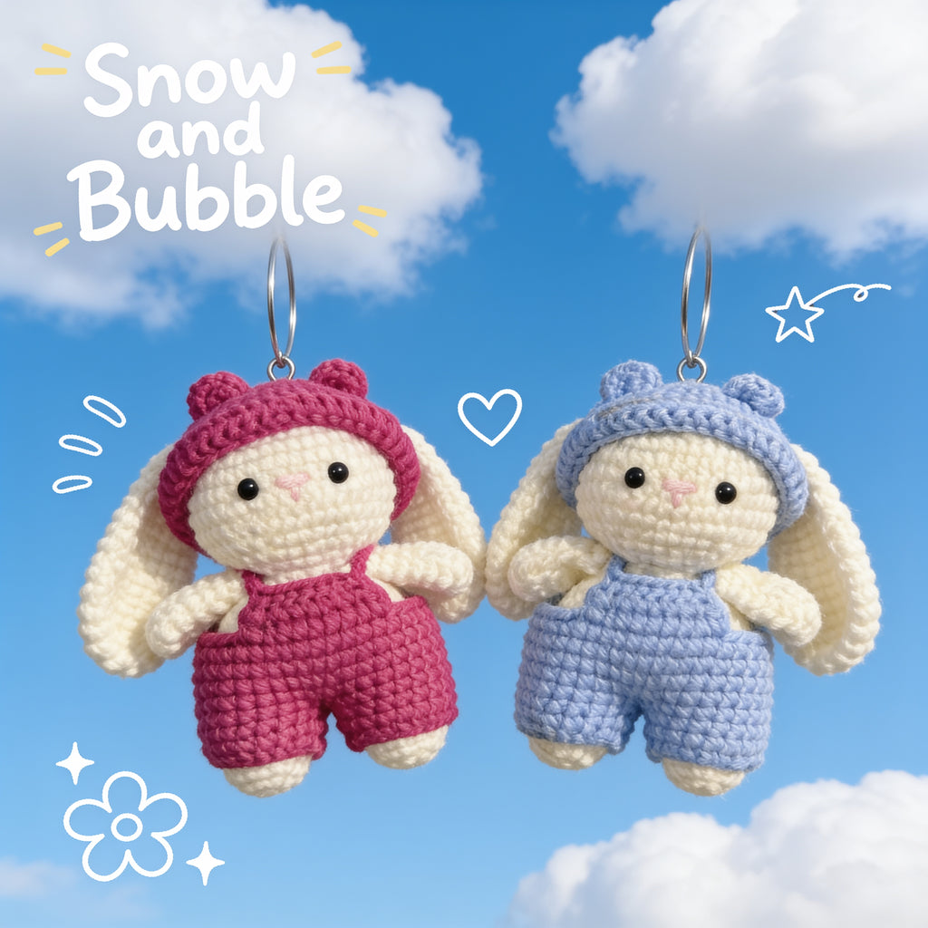 Two crocheted bunny charms hanging against a blue sky background with clouds. The left bunny wears a pink hat and pink romper, and the right bunny wears a light blue hat and blue romper.